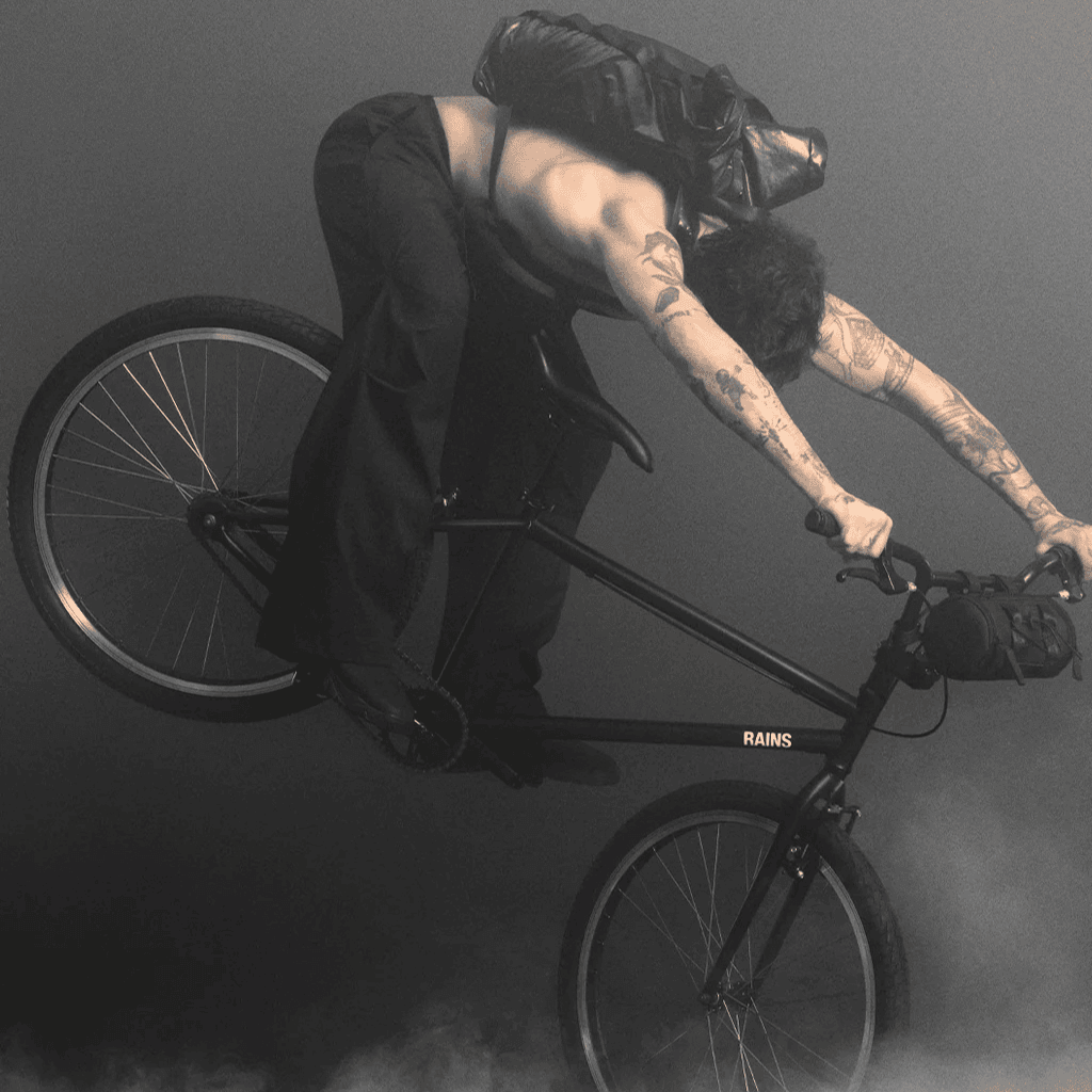 Bike rider performing a stunt on a mountain bike with tattoos, black clothing, and a backpack against a dark background.
