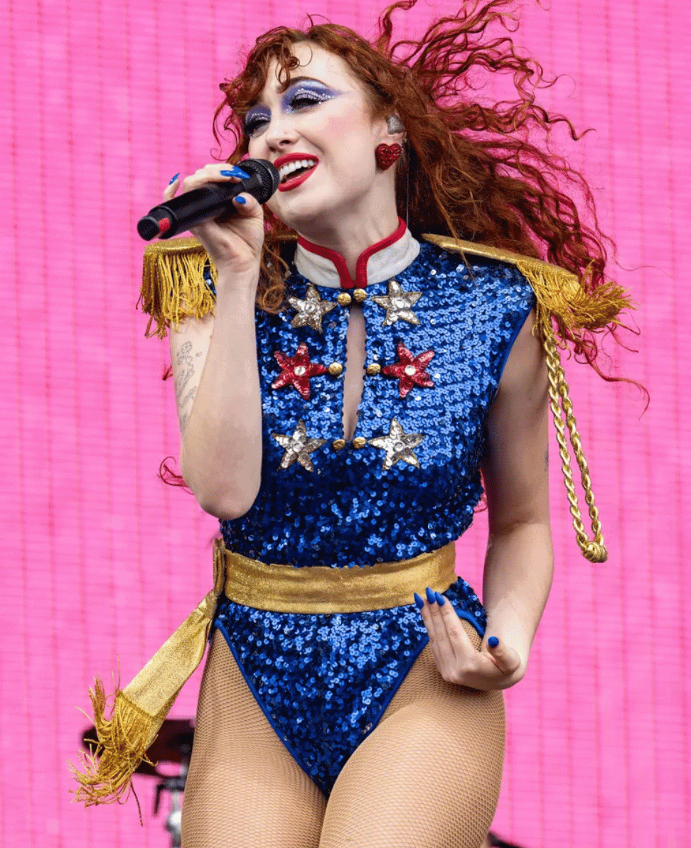 Vibrant performer with red curly hair and bold makeup singing on stage in a sequined patriotic costume.