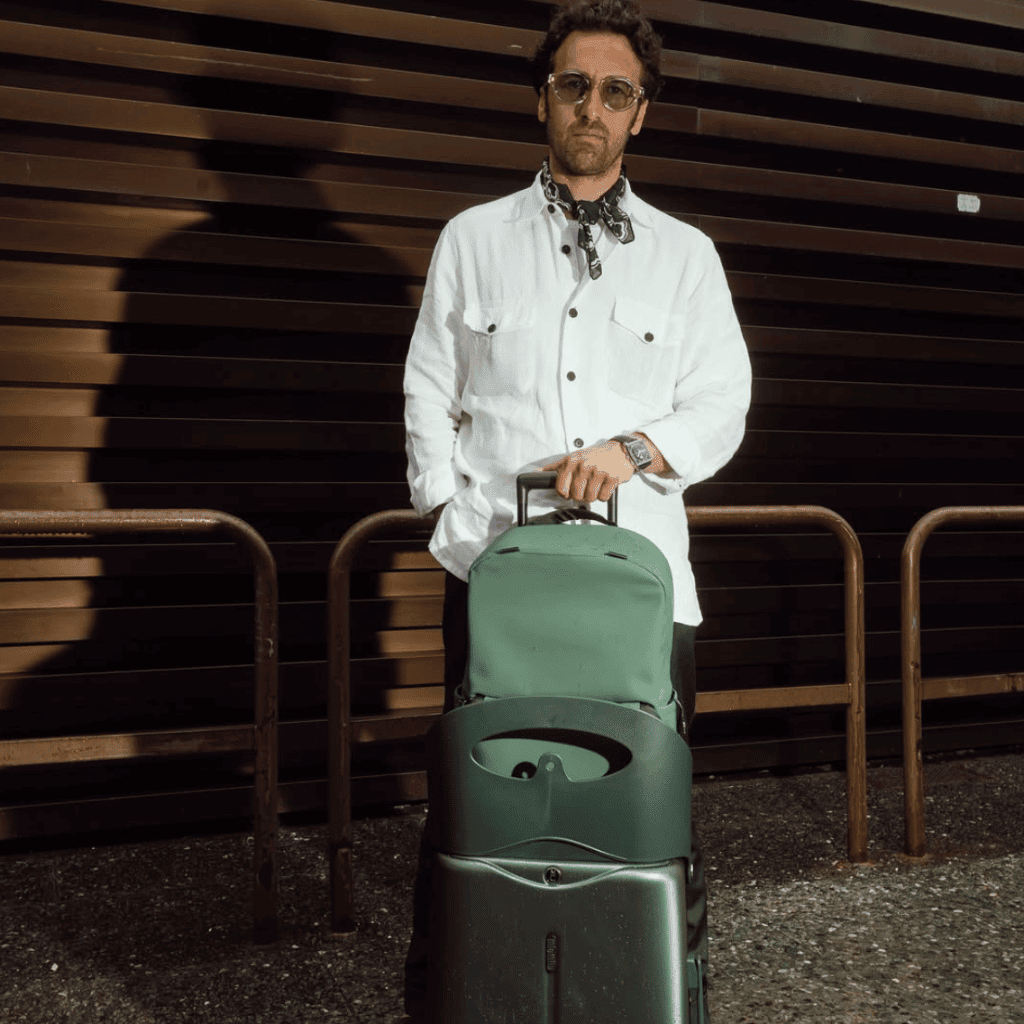 Suitcase with laptop bag on top, stylish man in white shirt and sunglasses, urban background, luggage for travel, modern travel accessories.