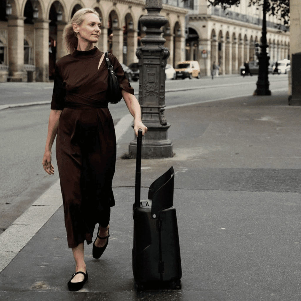 Travel-ready woman walking on city street with luggage and handbag, stylish urban travel outfit, modern cityscape background.
