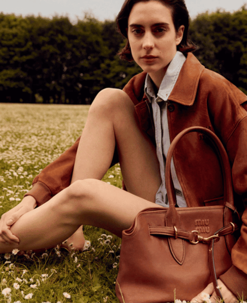 Handsome young woman sitting outdoors in field of flowers, casual style, brown leather bag, autumn clothing for fashion and lifestyle shot, lushcollective.com.