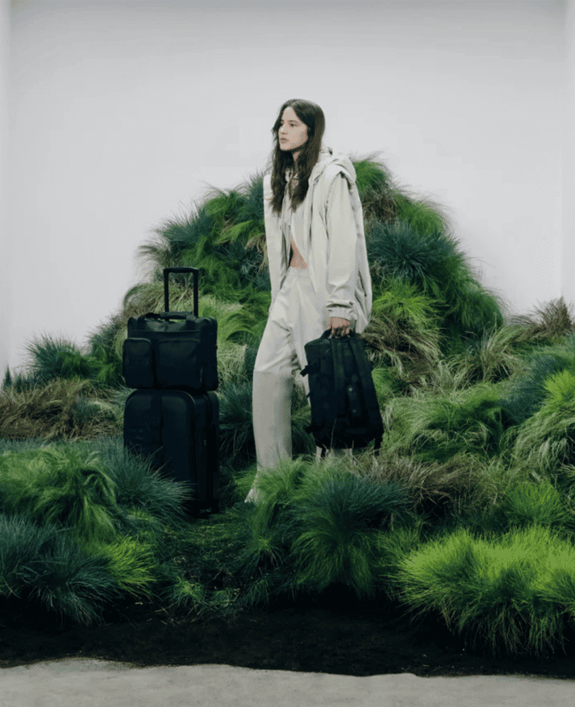 Black travel luggage with woman in beige outfit in a green moss landscape, modern luggage for travel and adventure, urban travel scene, Lush Collective inspiration, stylish travel accessories.