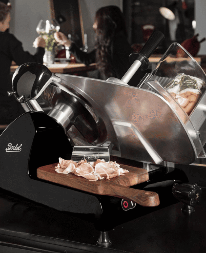 Slicer machine cutting chicken meat in a culinary setting with customers dining in the background.