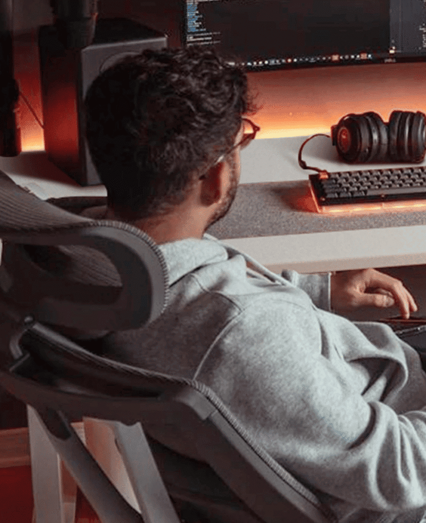 Relaxed man working at a modern desk with gaming equipment and warm ambient lighting.
