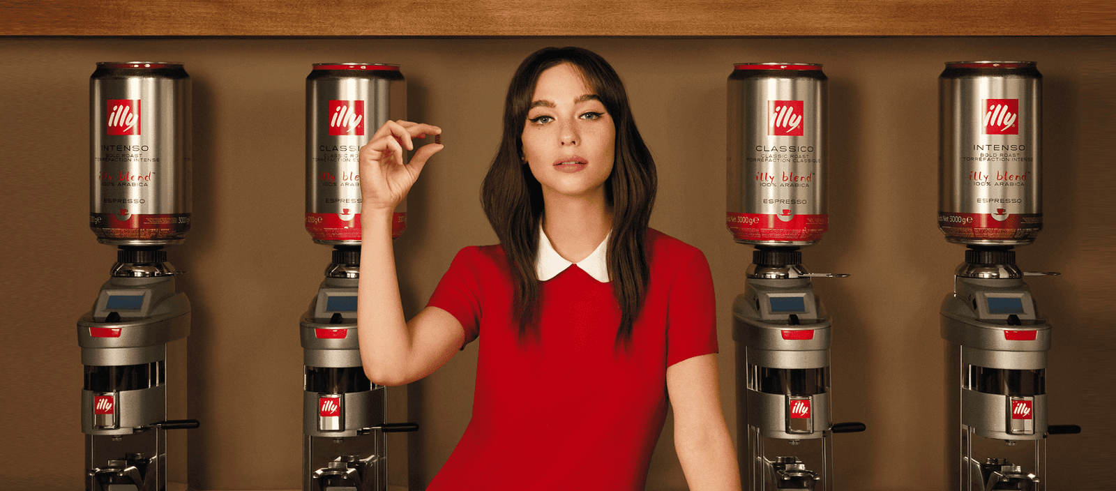 Illy coffee cans on coffee machines with a woman in a red dress, modern cafe setting, promoting premium espresso beverages.