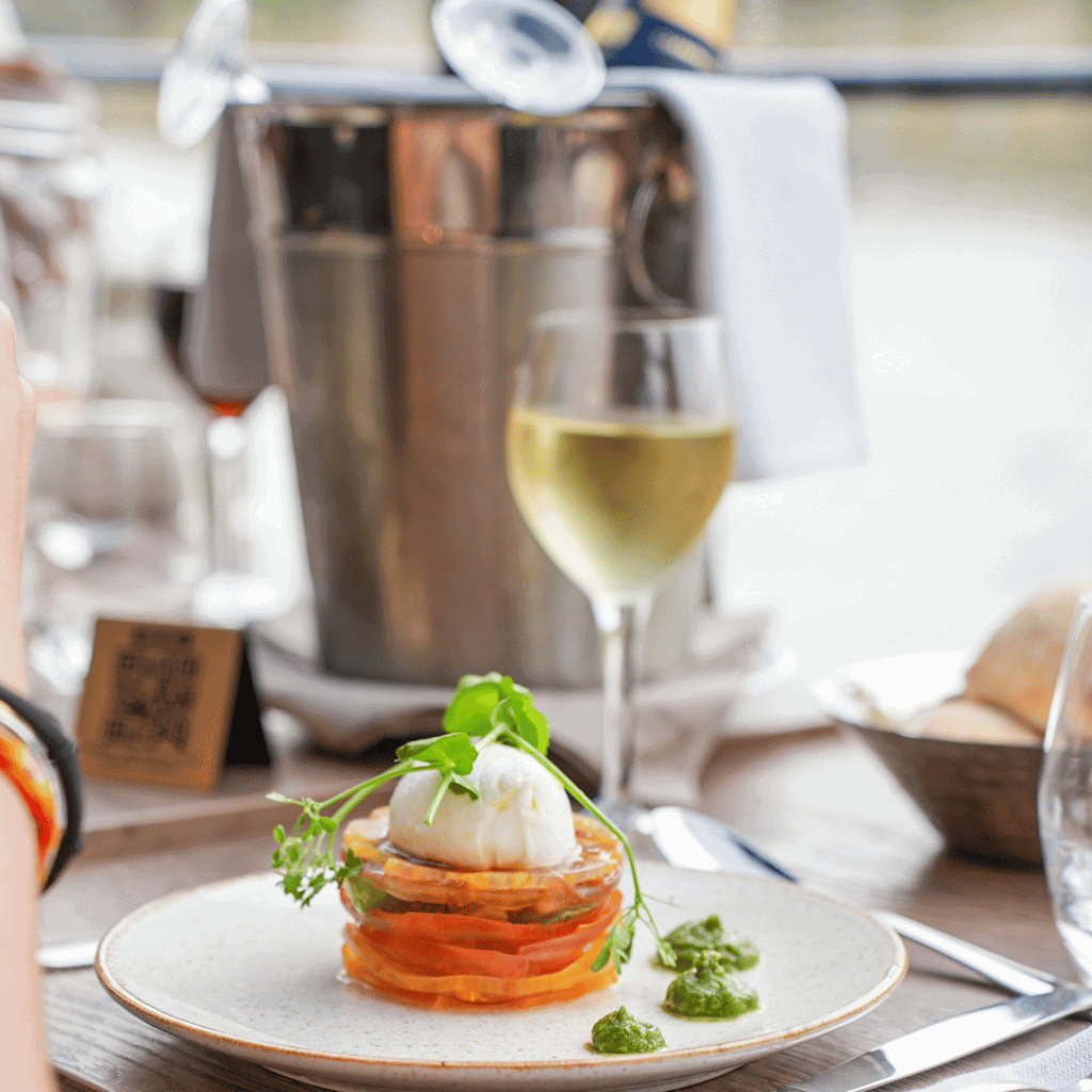 Fancy charcuterie plate with garnishes and fresh herbs, wine glass, and silverware on rustic table.