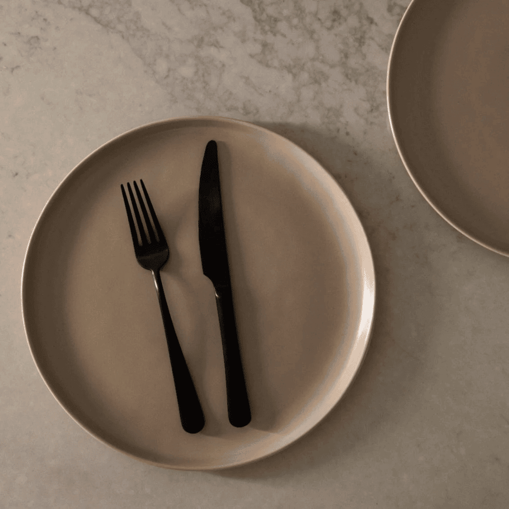 Minimalist beige plate with black fork and knife on a marble surface, modern tableware arrangement.
