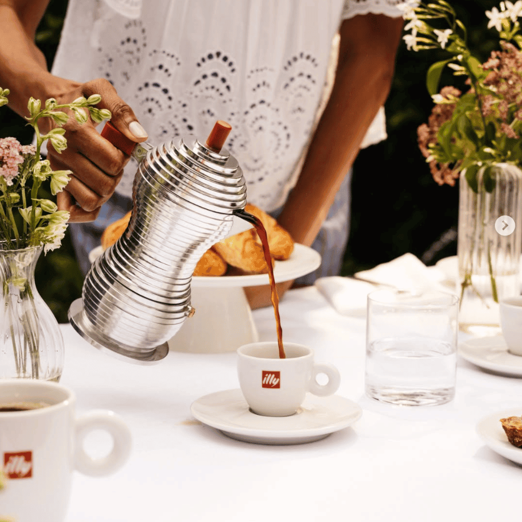 Hand pouring coffee from vintage teapot into a white cup at outdoor brunch scene.
