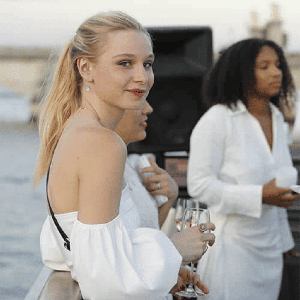Elegant women at a luxury outdoor event, enjoying drinks and socializing by the waterfront. High-end fashion and contemporary lifestyle scene.