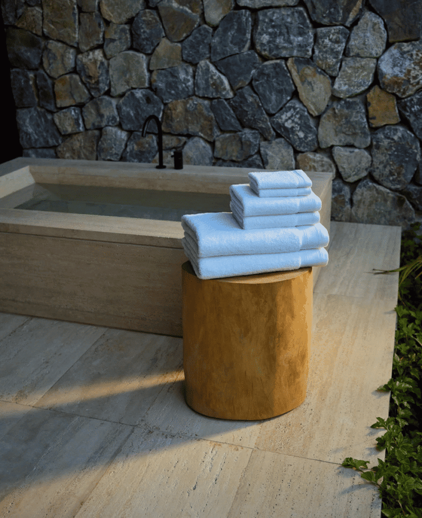 Lush Collective outdoor bathtub with fresh white towels on wooden stool, stone wall background, relaxing spa setting.