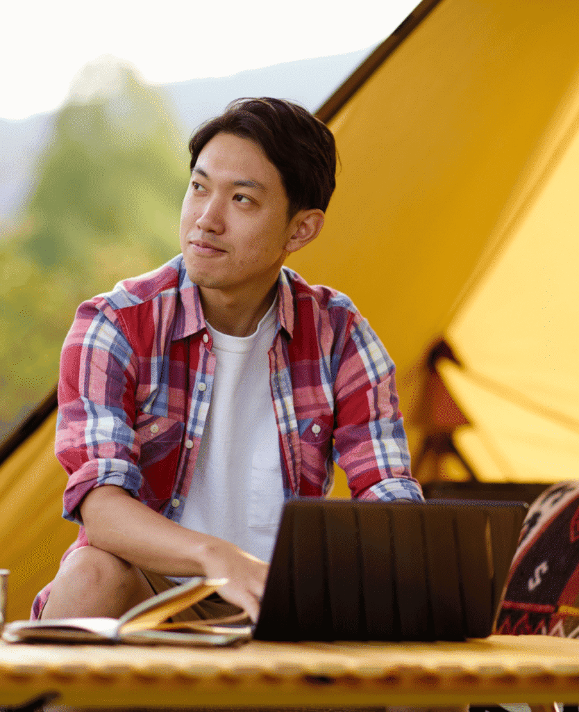 Relaxed man with a laptop at a camping tent, enjoying outdoor work or leisure in nature, lifestyle, adventure, outdoor camping, digital nomad.