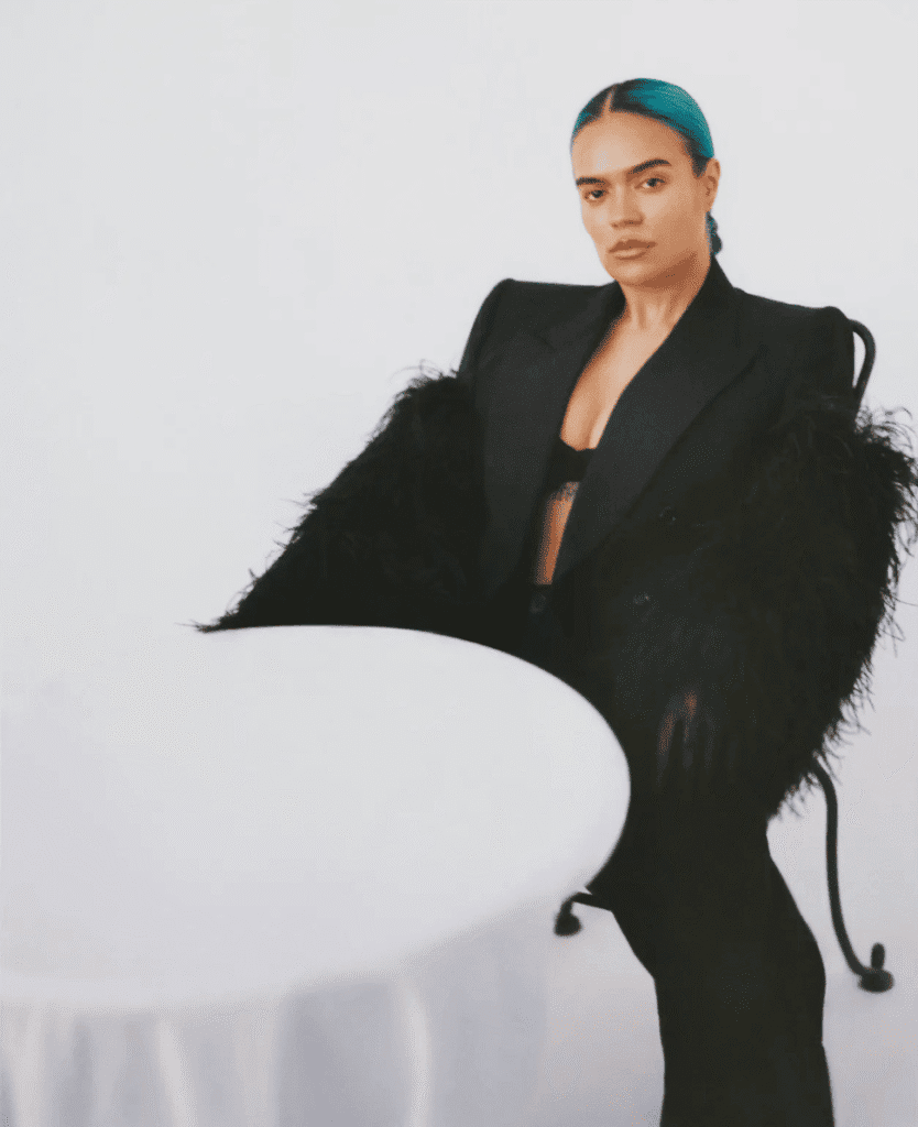 Bold woman with blue hair wearing a black blazer and feathered sleeves sitting at a white table.