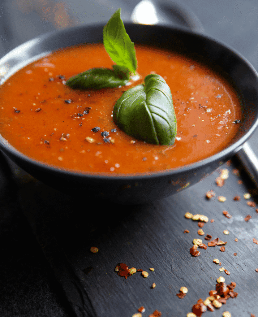 Savory tomato soup garnished with fresh basil leaves on black slate, spices, and chili flakes.